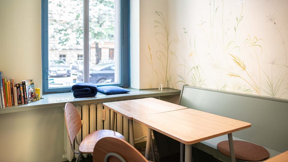 Sitzecke mit Holztisch vor einer handgemalten Gräser-Wandmalerei, Kinderbücher und Spiele auf der Fensterbank